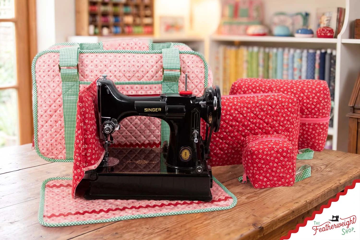 The Stitcher's Suitcase, a black Singer Featherweight 221 and the Feathered Friends organizers together on a wooden table in a bright sewing room lined with fabric.