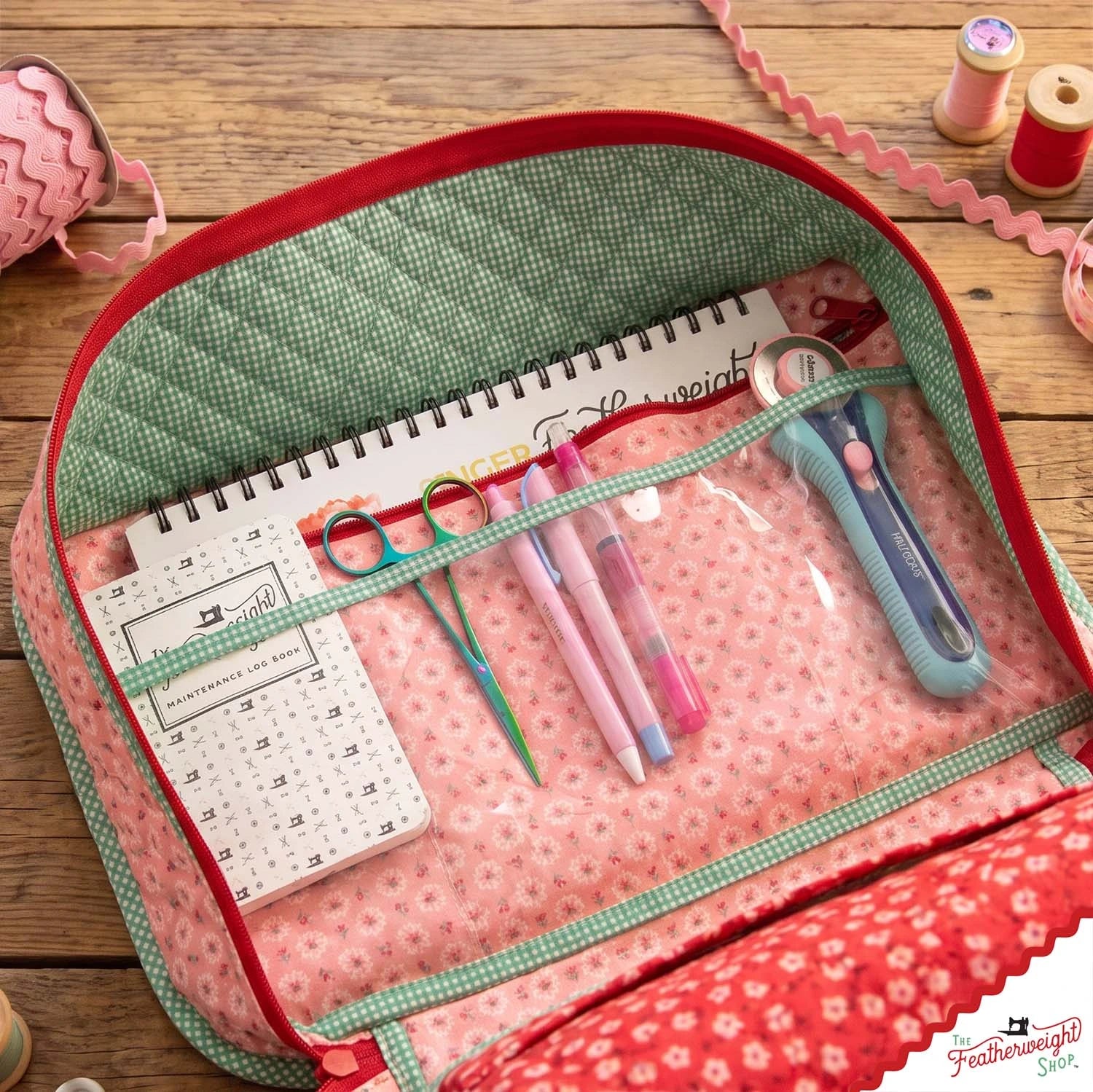 The clear vinyl notions pocket opened flat, holding a quilting ruler, scissors, marking pens and a rotary cutter on the pink medallion lining.