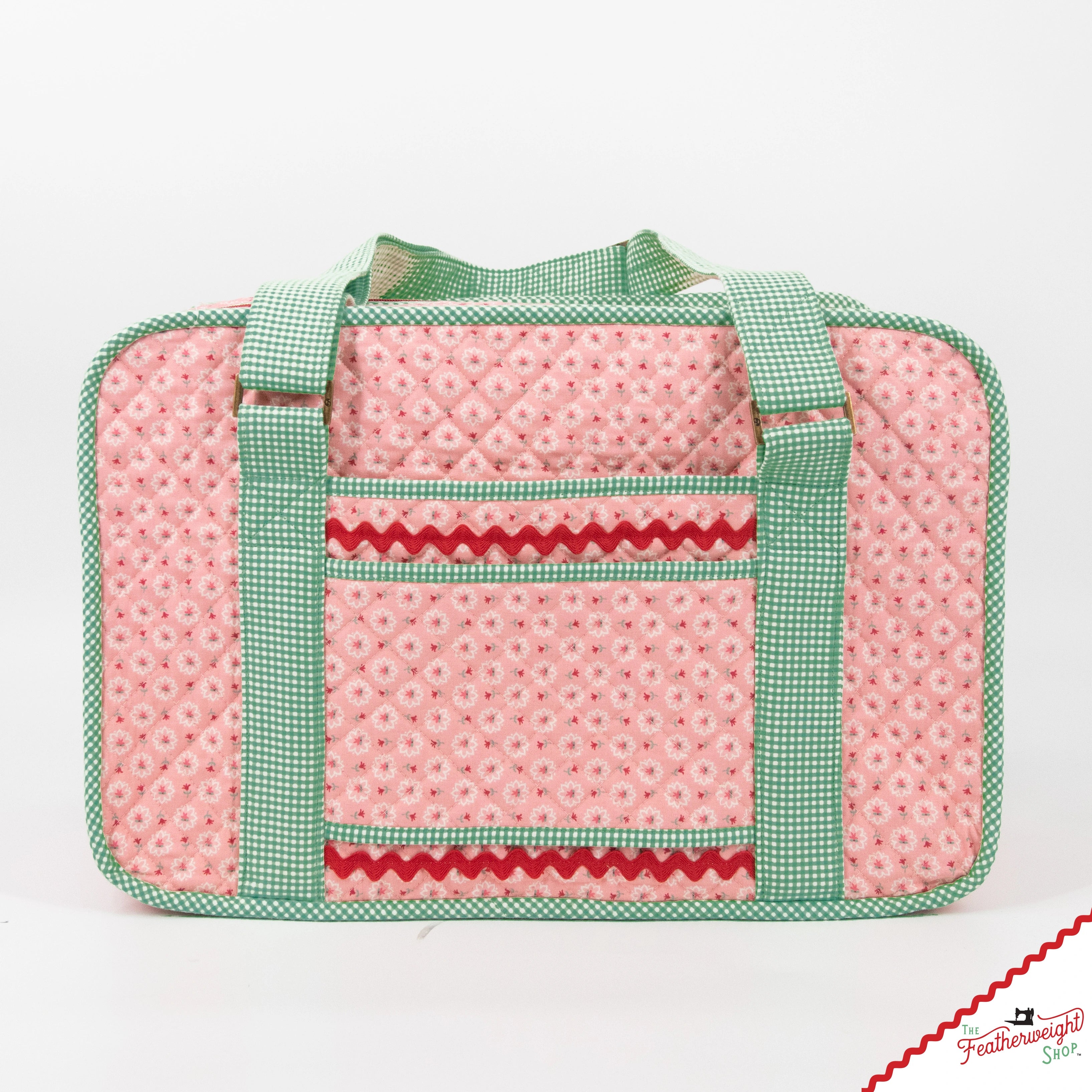 The finished Stitcher's Suitcase closed and seen from the front, sewn in pink Lori Holt medallion print with green gingham binding and handles and red ric rac trim across the front pocket.
