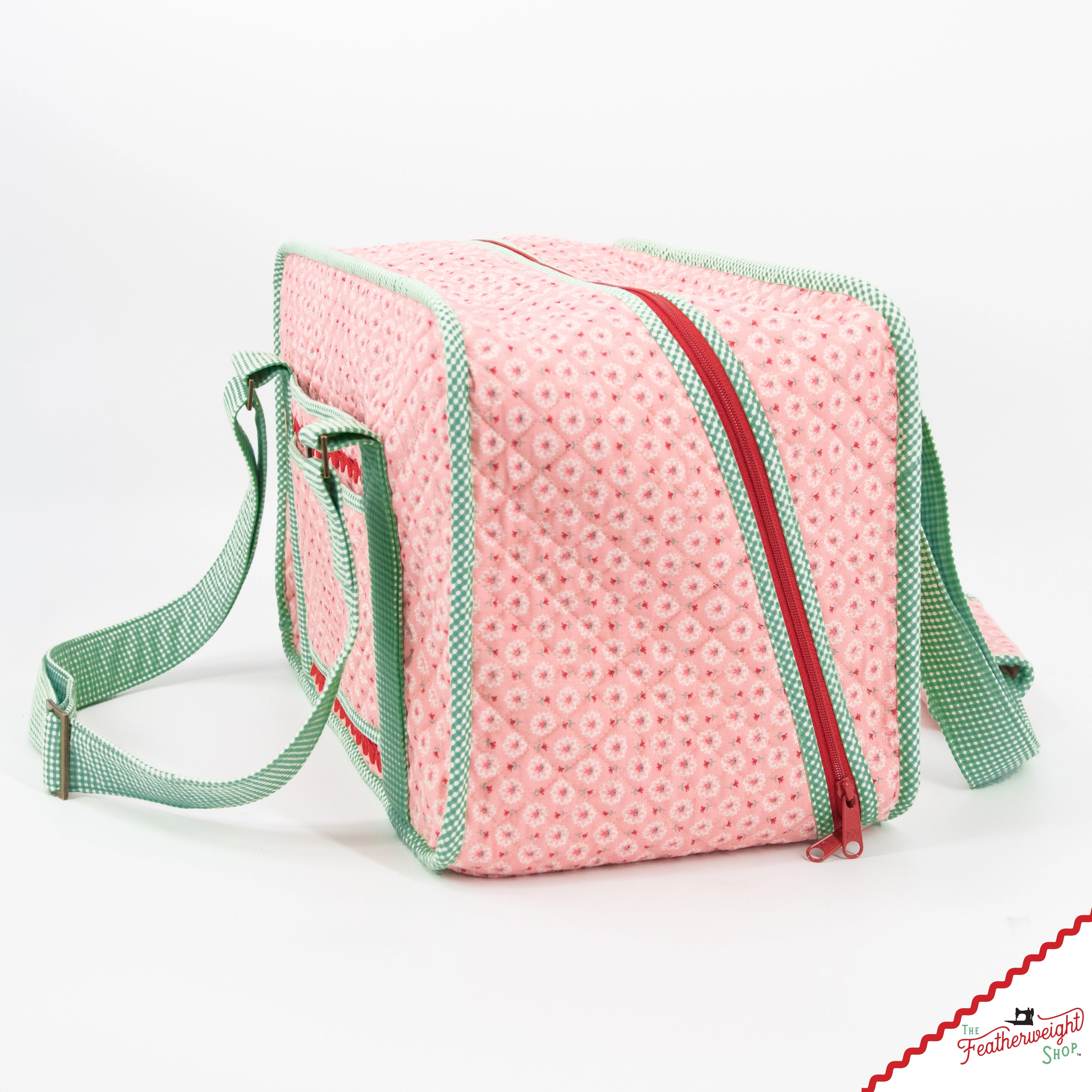 The Stitcher's Suitcase seen from the end, showing its boxy depth, the red zipper that runs around the top opening and the adjustable green gingham shoulder strap.