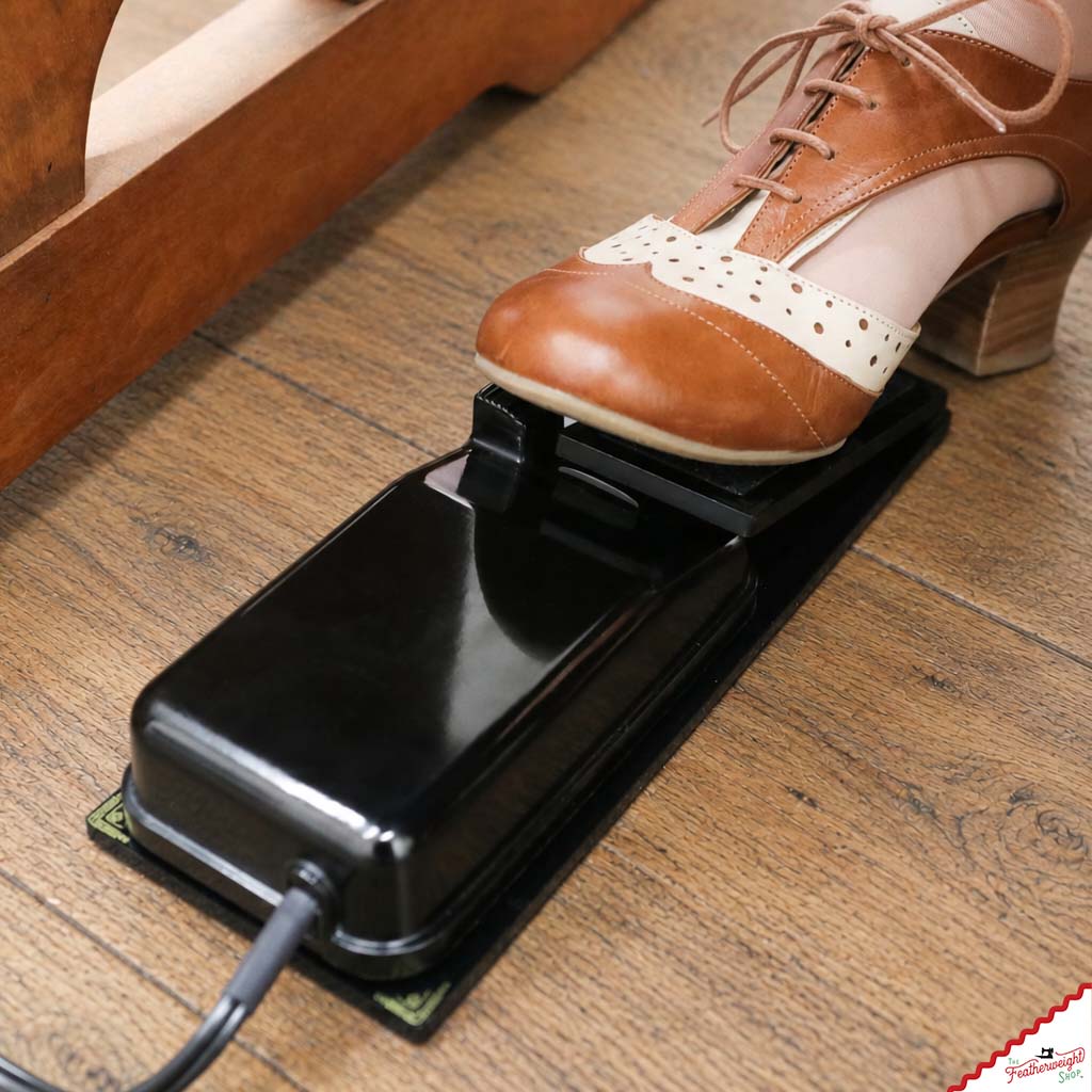 The Foot Controller Adaptor on a wood floor with the original foot pedal and its cord in the cradle, a shoe resting on the paddle.